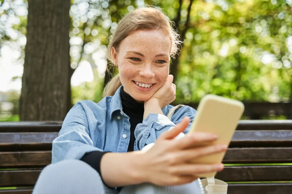 A woman sits on a bench, smiling while using her phone, reflecting joy and connection in her mental health journey