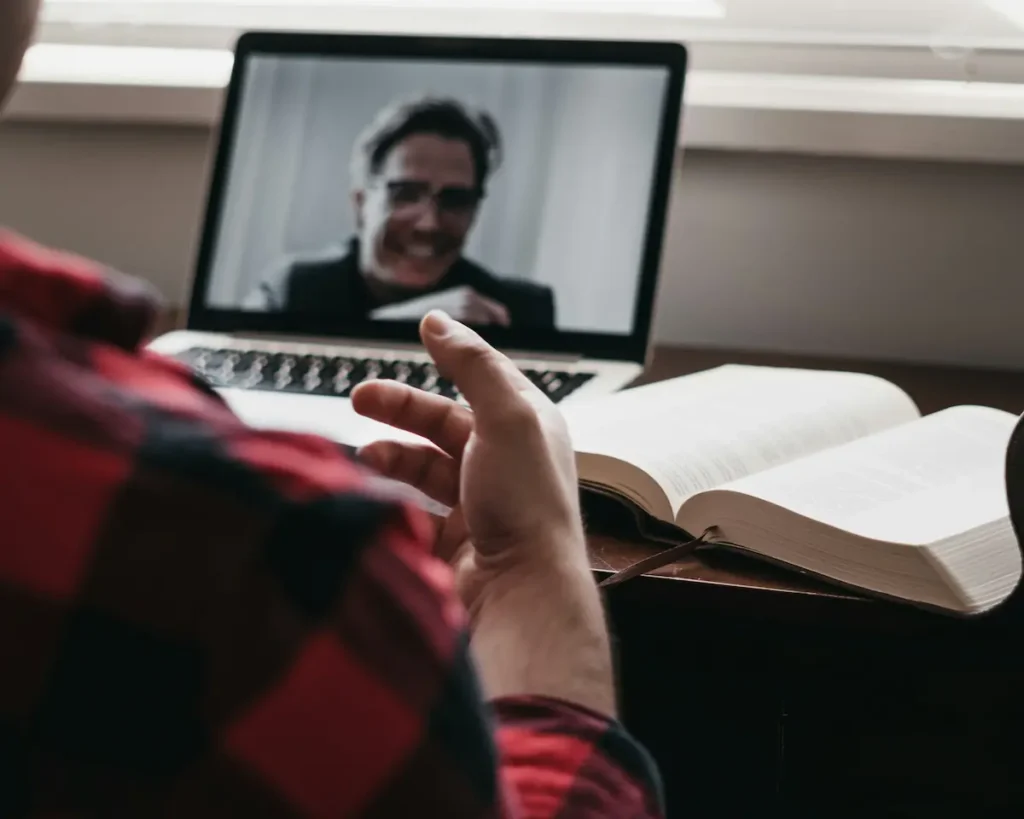 A man sits at a desk with a laptop and an open book, engaged in a gluco-keto online coaching session